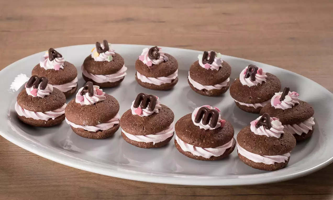Whoopies cioccolato scuro arrotondati con soffice crema rosa, decorati con riccioli di cioccolato su un piatto ovale candido