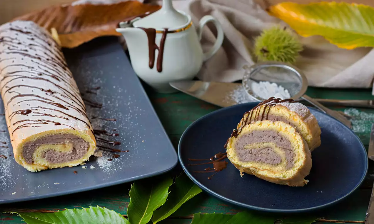 Kaštanová roláda světlé zlatavé barvy plněná jemným kaštanovým krémem, zdobená čokoládovou polevou na talíři vedle podzimních dekorací