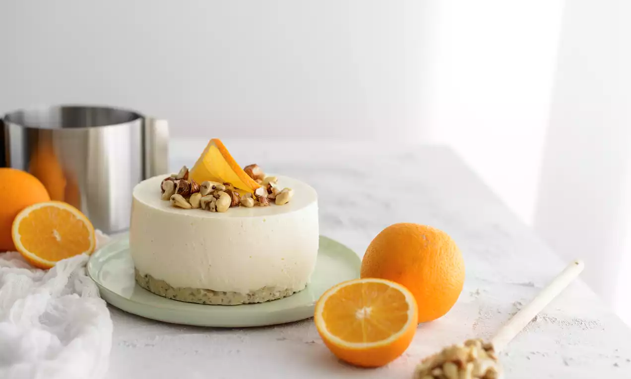 Helle Torte mit glatter Frischkäsecreme auf grünem Teller, garniert mit Nüssen und Orangenschnitzen, umgeben von frischen Orangen und einem Tortenring