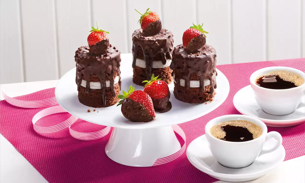 Zylindrische Schokotörtchen mit glänzender Knusperglasur, gefüllt mit weißer Creme, garniert mit Erdbeeren und Schokodekor auf einer Tortenplatte