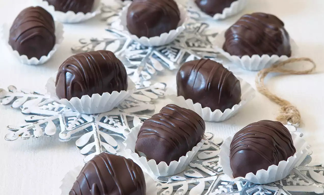 Švestkovo-marcipánové pralinky s lesklou tmavohnedou čokoládovou polevou, jemne zdobené prúžkami, uložené v bielych košíčkoch na striebristej vločke