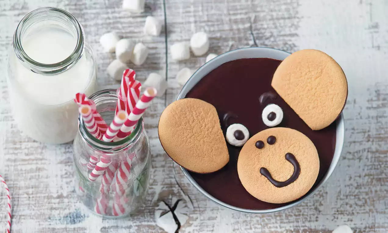 Čokoládový krém s hladkým povrchom v miske, ozdobený dvomi svetlohnedými sušienkami a čokoládovou tvárou v tvare usmievajúcej sa opičky