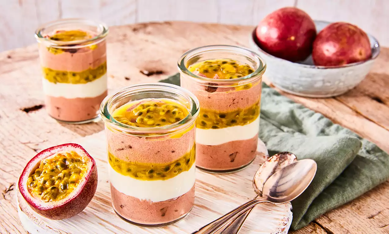 Schichtdessert aus brauner Schokomousse, weißer Creme und gelber Maracuja, serviert in Gläsern auf Holzbrett mit Löffeln und frischer Maracuja