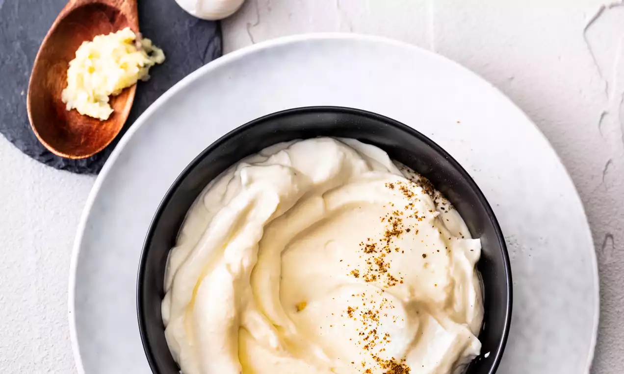 Creamy white aioli with a smooth texture swirled in a black bowl on a white plate, garnished with golden spices and garlic