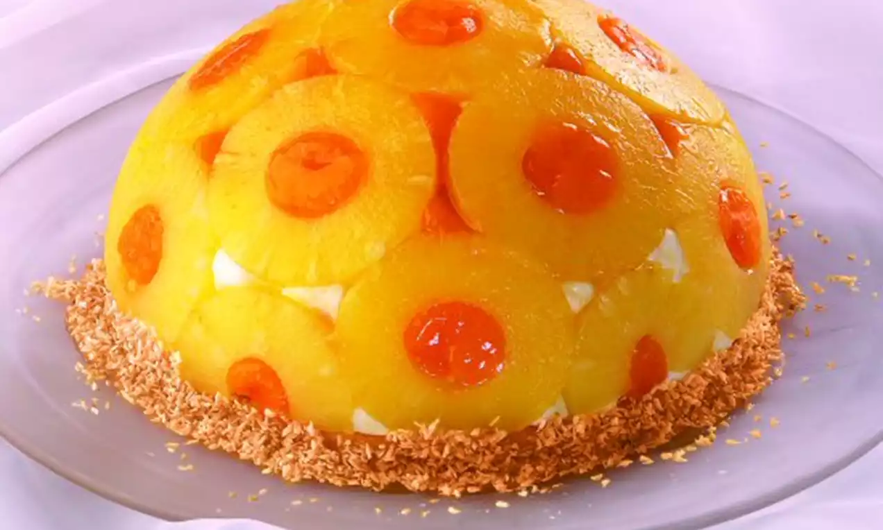 Kuppelförmige Torte aus goldgelben Ananasscheiben, verziert mit roten Kirschen auf Kokosraspeln, präsentiert auf einer transparenten Platte