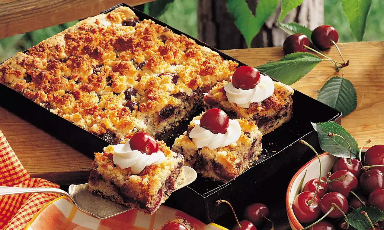 Saftiger Kirsch-Kokoskuchen mit goldbrauner Streuseldecke, Stücke mit Sahnehäubchen und Kirsche garniert, auf schwarzem Blech serviert