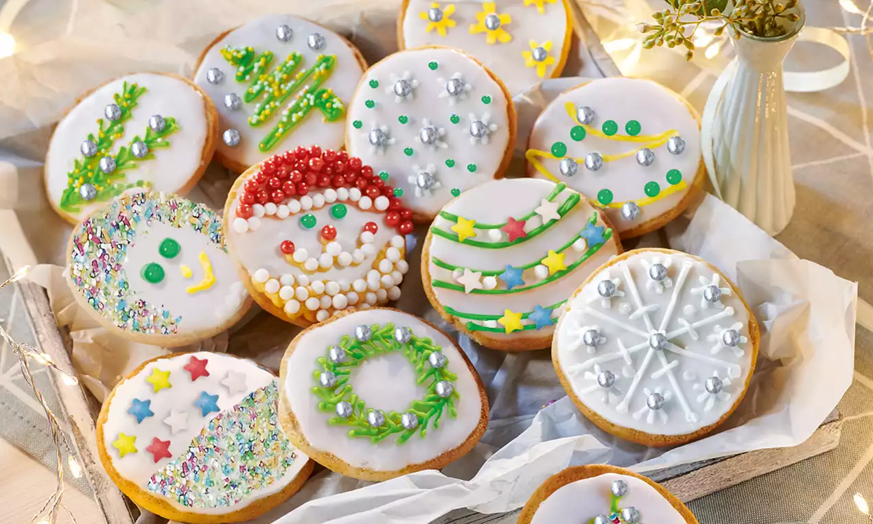 Beyaz glazürle kaplanmış rengarenk detaylarla süslenmiş yılbaşı temalı kurabiyeler ahşap kutuda ışıklarla çevrili şekilde sunulmuş