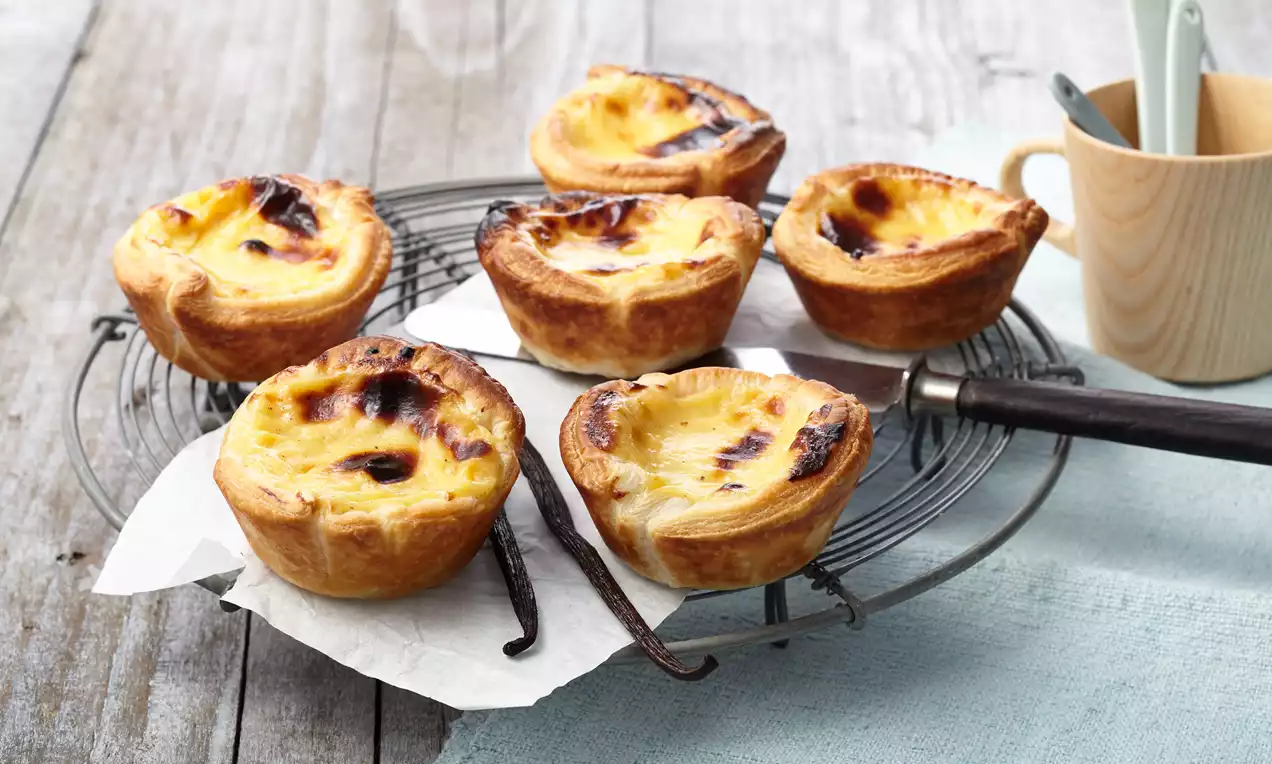 Kleine gouden pasteitjes met geblakerde bovenkant op een metalen rooster met bakpapier, naast een houten kopje met lepels op een blauw doek