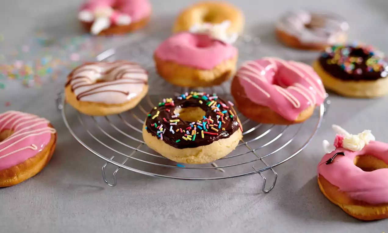 Goudbruine donuts met glazuur in roze, wit en chocolade, kleurrijk bestrooid met sprinkles, gerangschikt op een metalen rooster