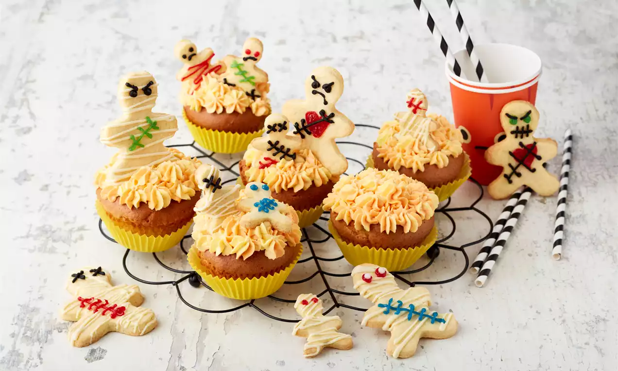Halloween-Cupcakes mit karamellbraunen Böden, cremigem weiß-orange Quark-Topping, Zuckerguss-Flammen und Mürbeteig-Puppen sowie Mumien