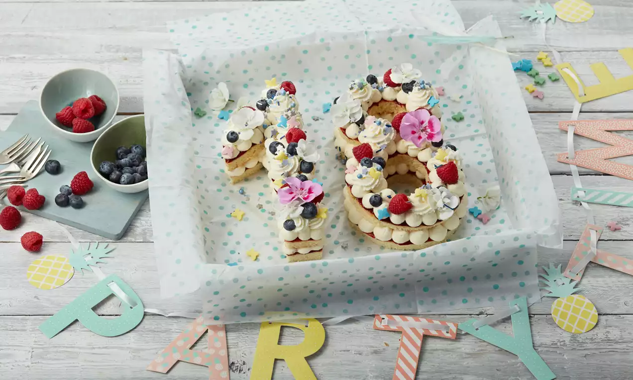 Chiffres 18 en pâte dorée superposée garnis de crème blanche, fruits rouges, fleurs roses et décorations colorées sur papier à pois verts
