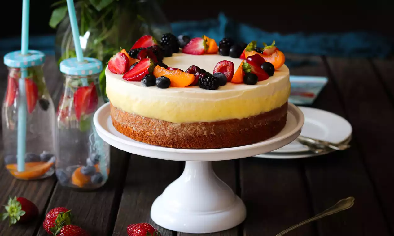 Svetložltá ovocná torta na svetlom podstavci s vrstvami krémovej plnky, ozdobená čerstvými jahodami, malinami, černicami a marhuľami