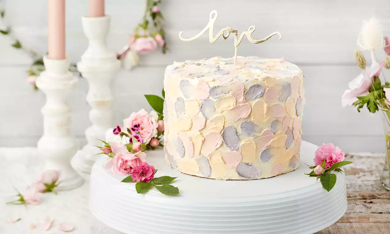 Runde kleine Torte mit pastellfarbener, floral gemusterter Buttercreme und goldfarbenem Schriftzug auf weißer Tortenplatte mit Rosen dekoriert