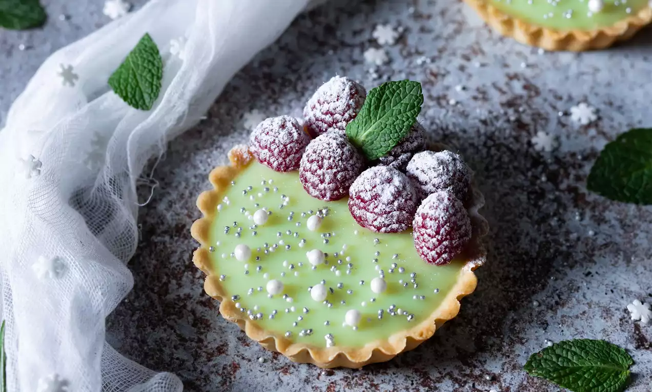 Tartaletka s hladkým světle zeleným krémem, zdobená malinami posypanými moučkovým cukrem, stříbřitými perlami a lístkem máty na rustikálním podkladu
