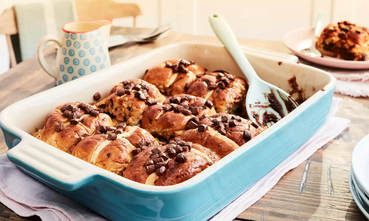 Golden hot cross buns nestled in a large blue baking dish, topped with glossy melted chocolate chips alongside a pale green spoon