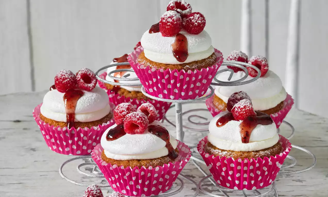 Pavlova cupcakes v růžových papírových košíčcích, zdobené bílým sněhovým krémem, čerstvými malinami a jahodovým sirupem na kovovém stojanu