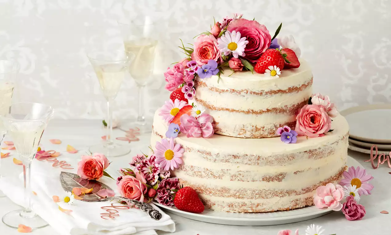 Zweistöckige Naked Cake mit heller Creme und sichtbarem Biskuit, dekoriert mit Rosen, Erdbeeren und bunten Blumen auf weißer Platte