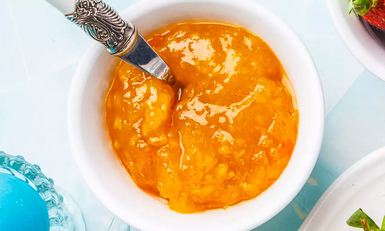 Oranje confituur met glanzende, gladde textuur in een wit schaaltje, geserveerd met een zilverkleurige lepel tegen een lichtblauwe achtergrond