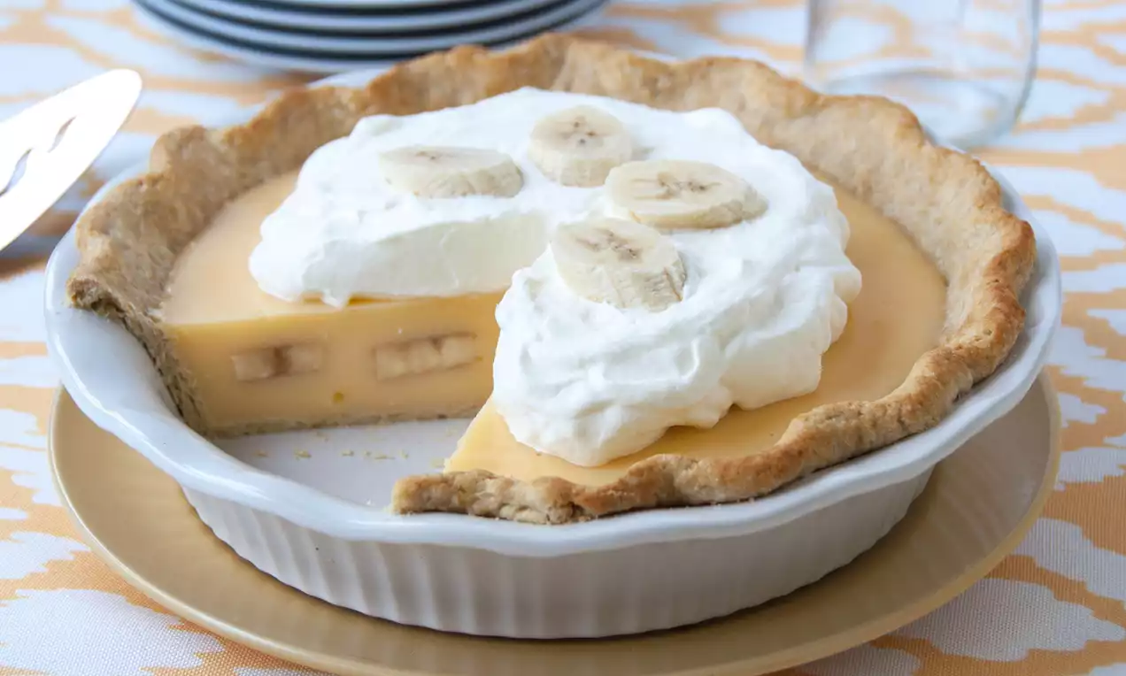 Tarte à la banane dans une pâte dorée, garnie de crème fouettée blanche et de rondelles de banane, présentée dans un plat à tarte blanc