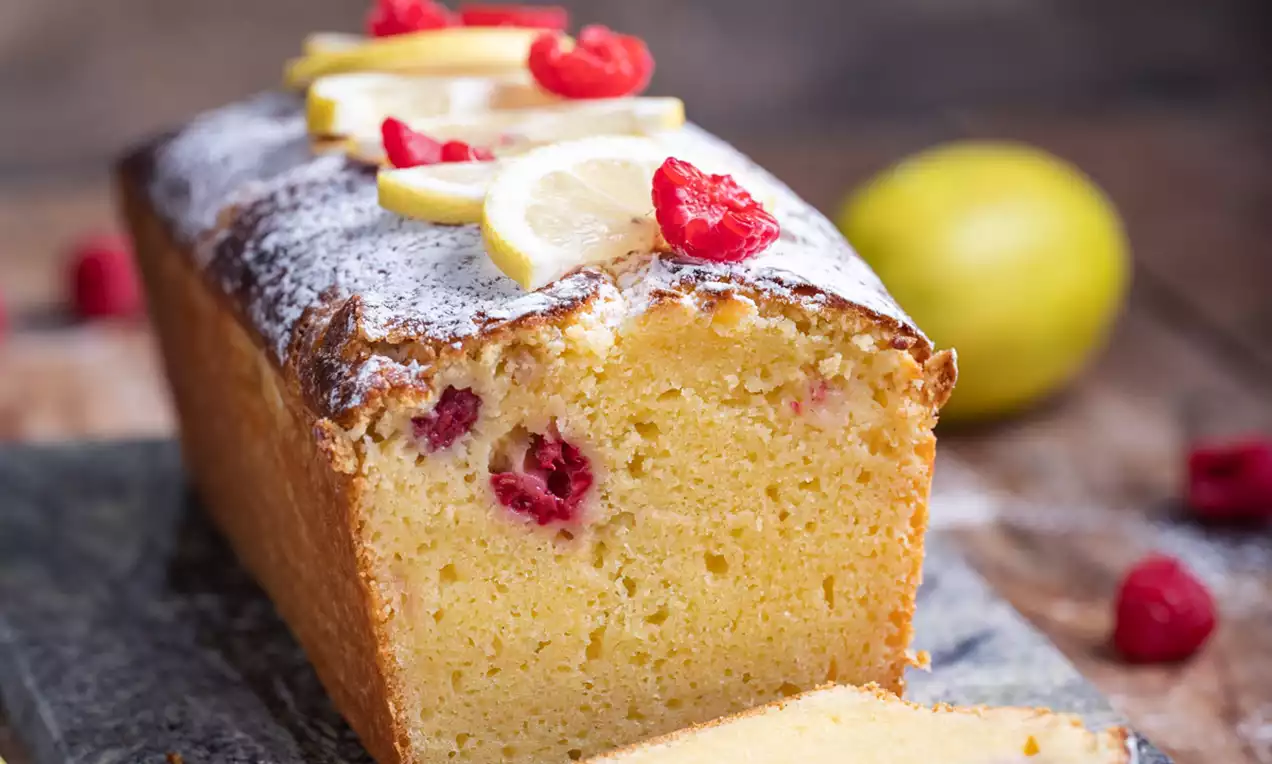 Duguljasti kolač svijetložute boje s malinama i limunovima, prekriven bijelim šećerom u prahu, na tamnoj drvenoj podlozi
