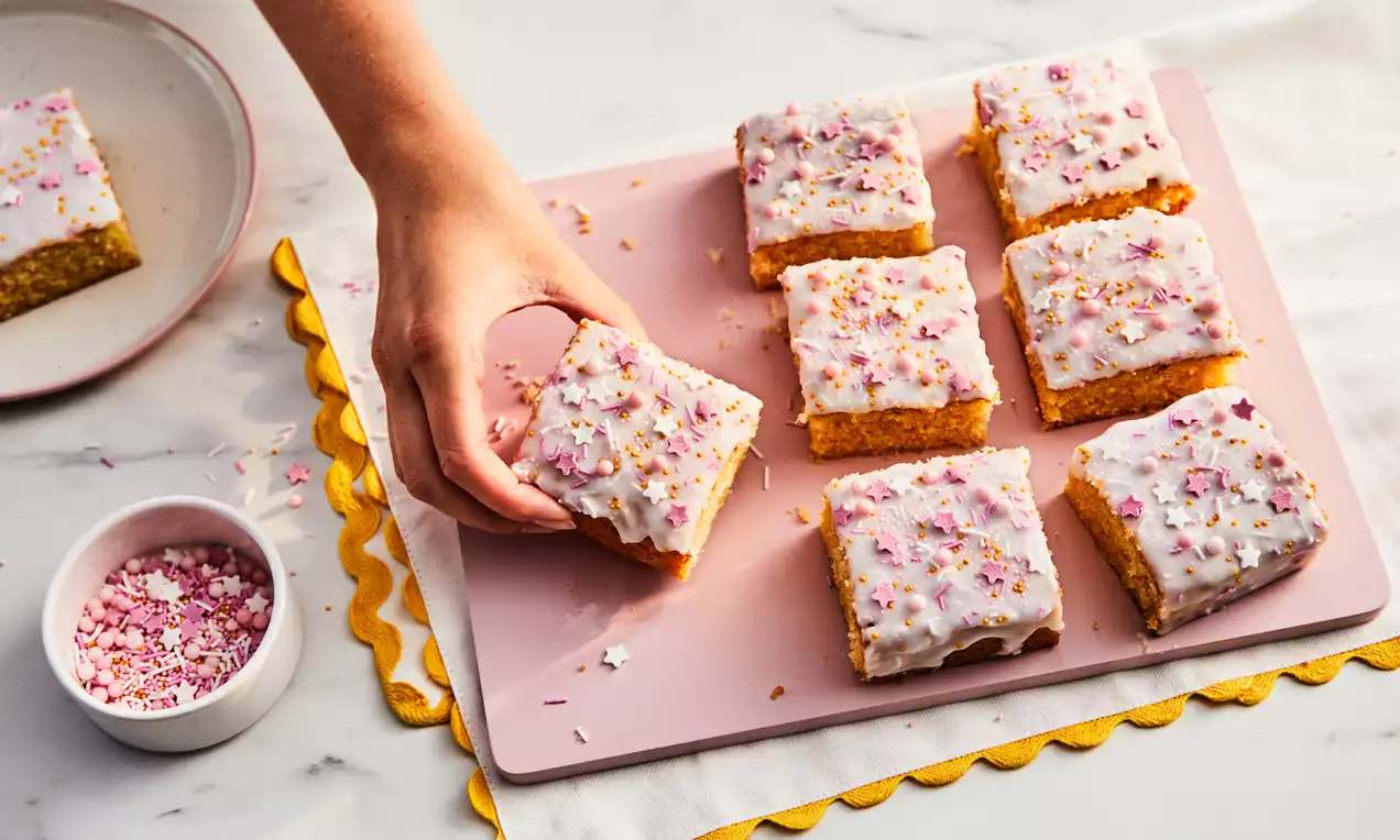 Golden vanilla sponge squares covered in pale pink icing and sprinkled with colourful sugar shapes on a pink board with a hand reaching