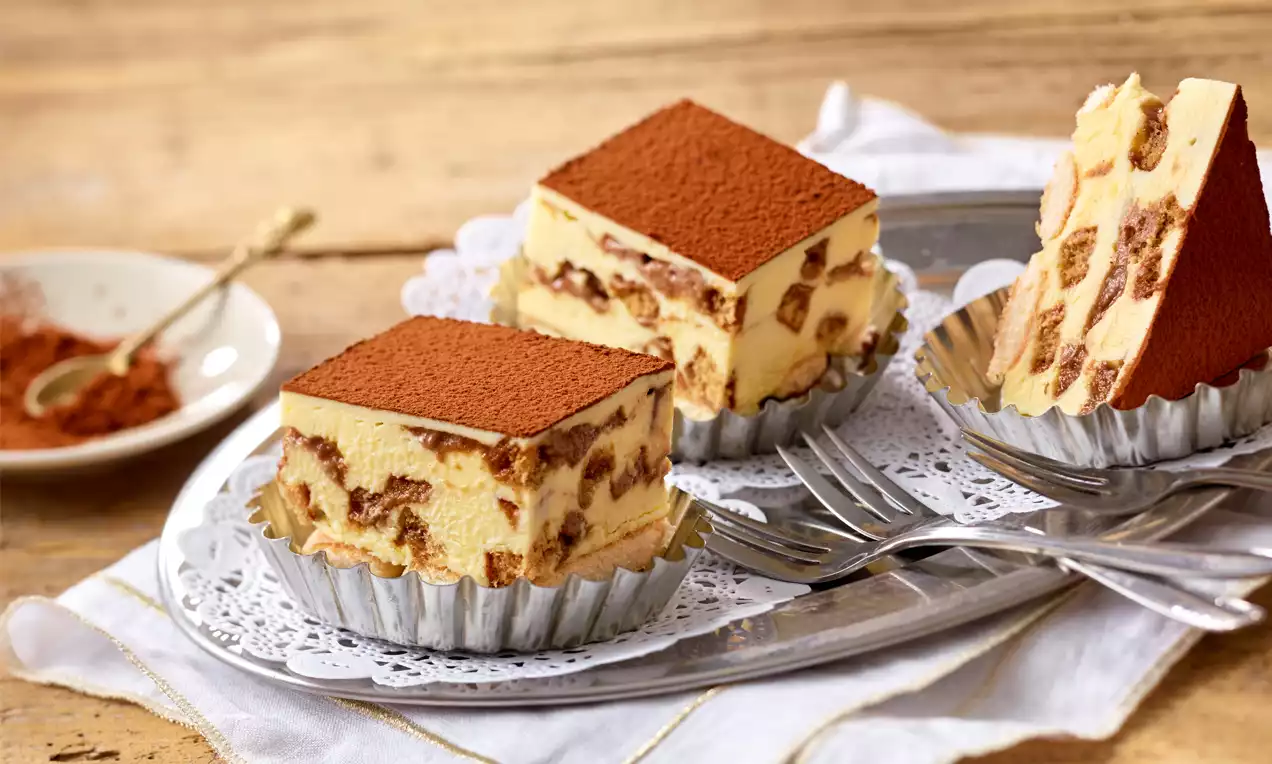 Drei Stück Lebkuchen-Tiramisu mit gelber Creme und braunen Lebkuchenstücken, oben bestäubt mit Kakao, serviert auf Silberförmchen mit Gabeln