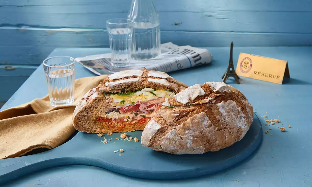 Rundes, goldbraunes Brot mit Mehl bestäubt, mittig aufgeschnitten, gefüllt mit bunten Schichten Gemüse und Belag auf blauem Holzbrett