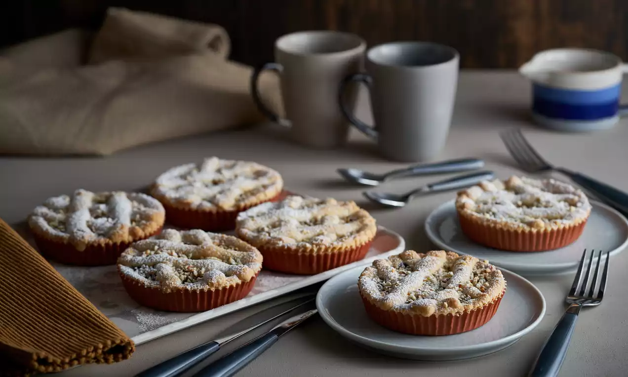 Crostatine dorate con intreccio di pasta friabile, spolverate di zucchero a velo e servite su piatti bianchi accanto a tazze dai toni neutri