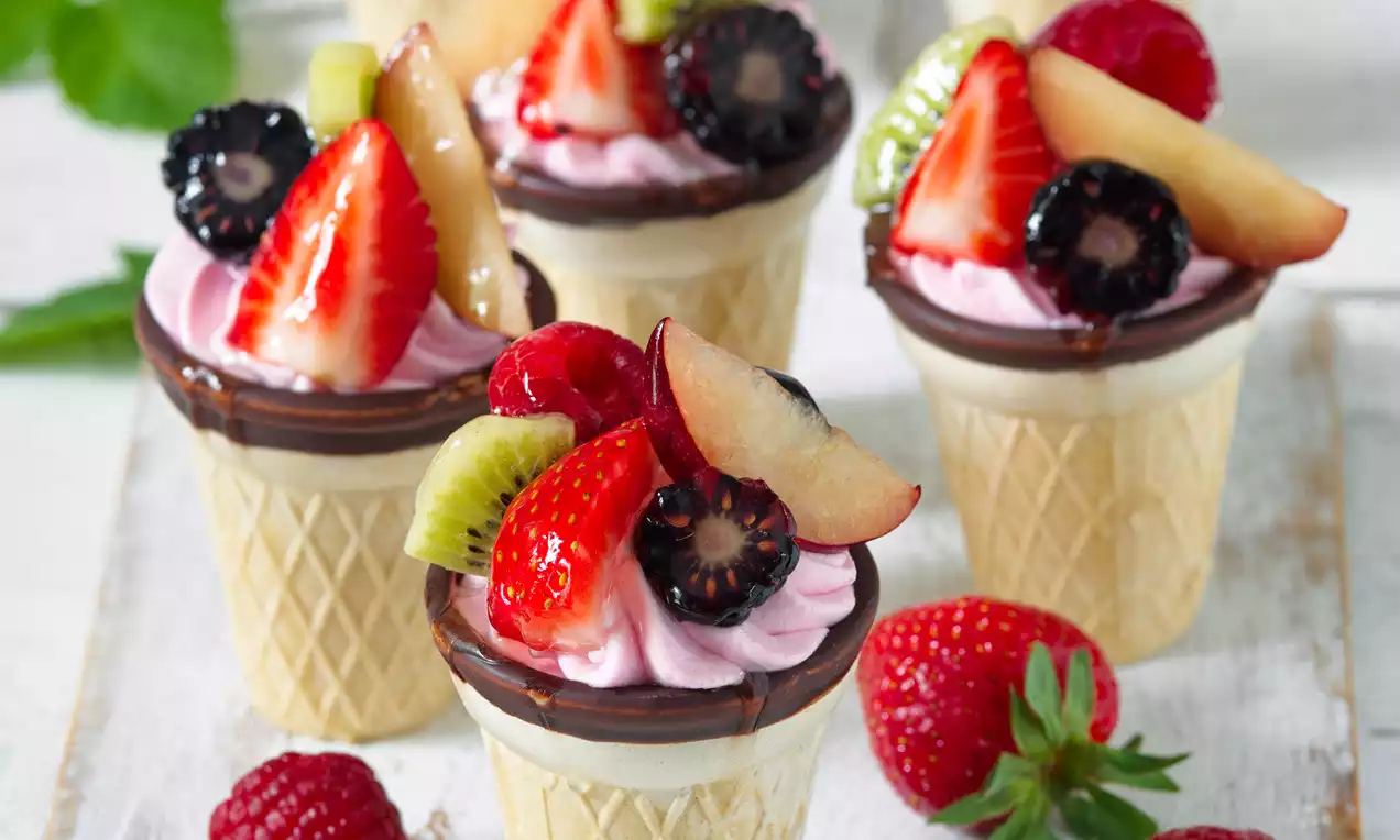 Zmrzle krémovo-ružové kornútky ozdobené čerstvým ovocím, na farebnej čokoládovej vrstve v krehkom vaflovom kuželi na svetlom podklade