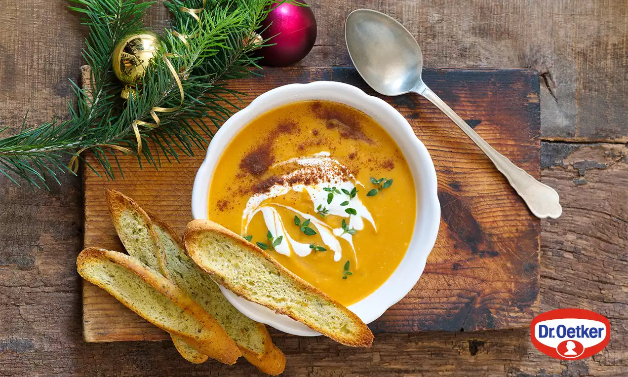 Krémová oranžová polievka v bielej miske, ozdobená šľahačkou, bylinkami a škoricou, podávaná s chrumkavými toastami na drevenom podnose