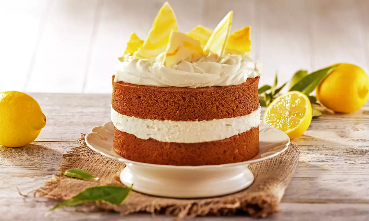 Torta alta a strati con soffice crema bianca al centro, decorata con riccioli di cioccolato bianco e limone, su piatto chiaro con limoni freschi accanto
