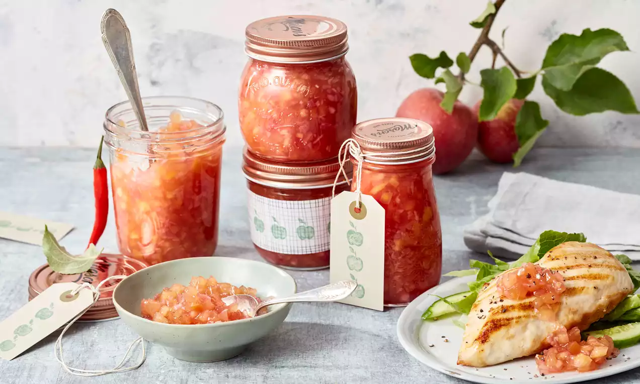 Apfelchutney in Gläsern mit Kupferdeckeln neben einem Teller mit gegrilltem Hähnchenfilet, frischen Äpfeln und einem Zweig als Deko