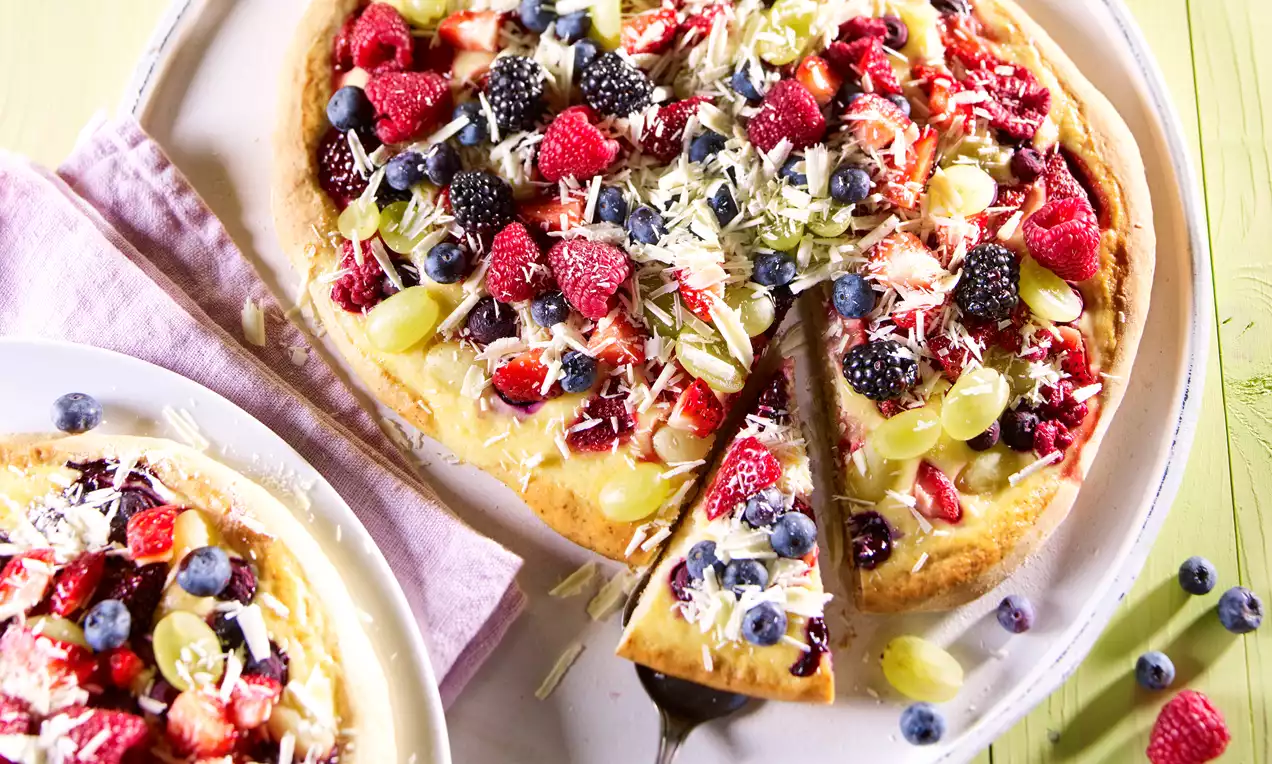 Helle Dessert-Pizza mit bunten Früchten wie Beeren und Trauben, geraspelter weißer Schokolade und einem herausgehobenen Stück auf hellen Tellern