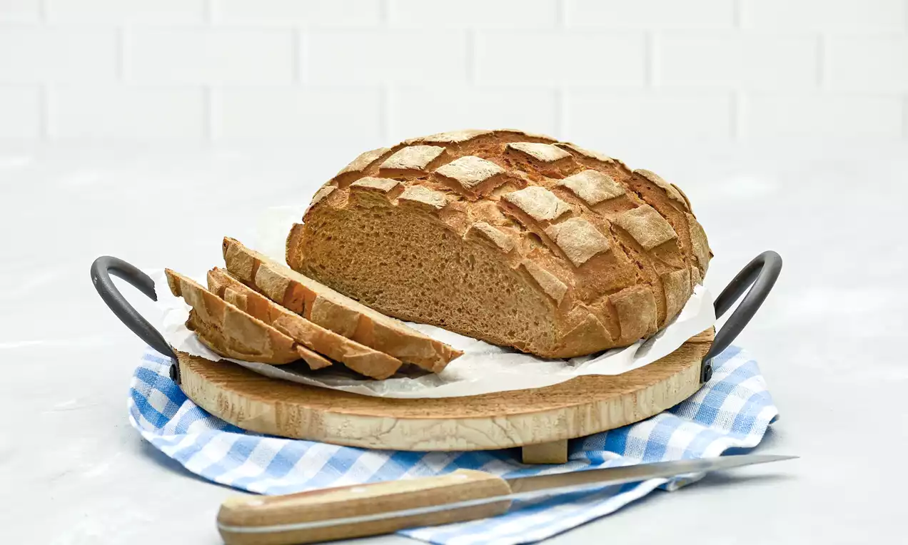 Goldbraunes Krustenbrot mit rautenförmigen Einschnitten, halb aufgeschnitten, auf Holzbrett mit weiß-blauem Tuch und Messer drapiert