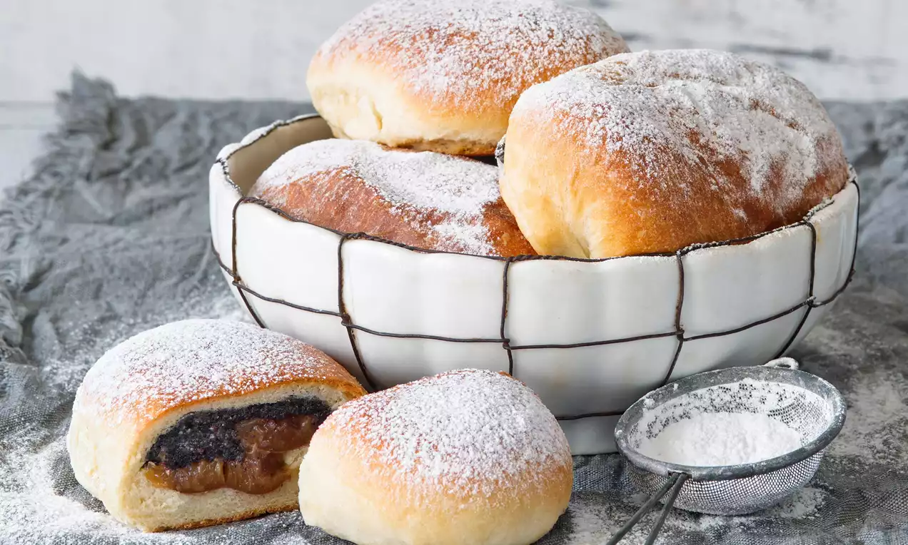 Slivkové buchty zlatohnedé a nadýchané, posypané práškovým cukrom, uložené v drôtikovanej miske na látkovom obruse vedľa sitka s cukrom