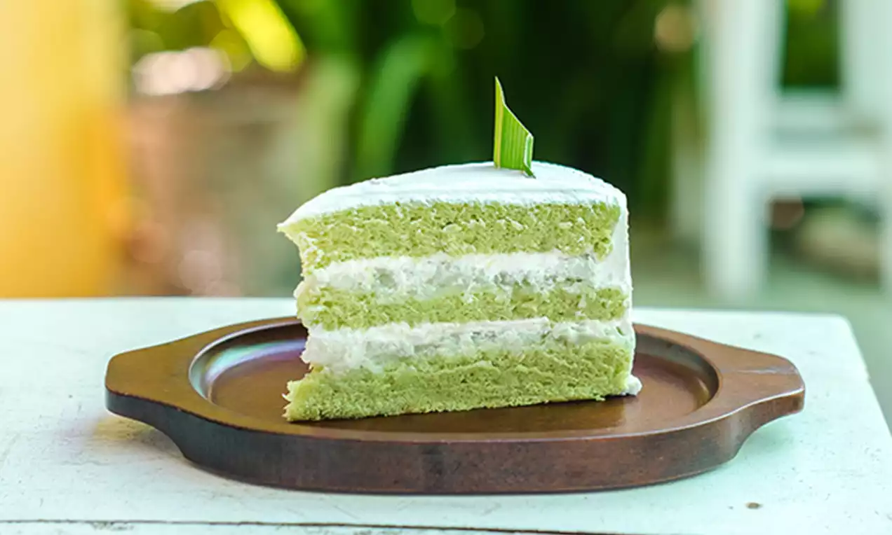 Layered slice of pale green pandan cake with fluffy white cream filling, topped with white icing and a single pandan leaf