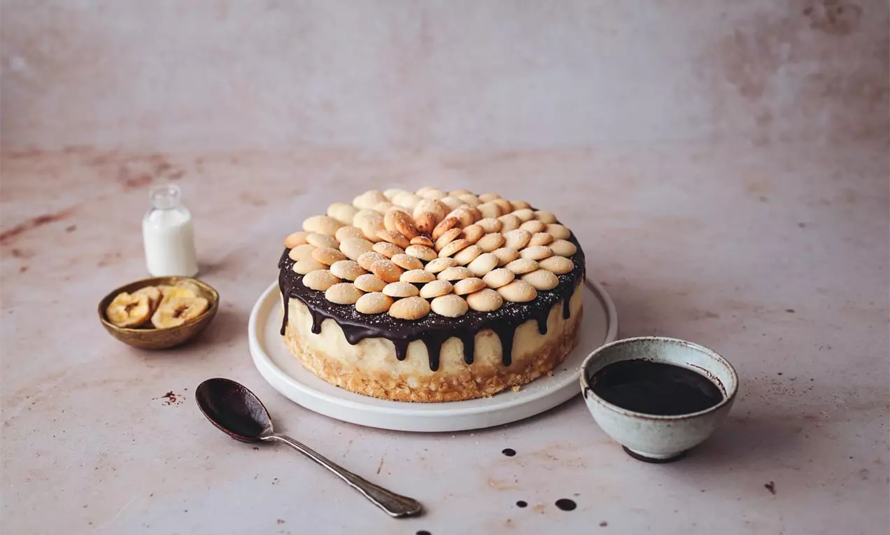 Okruhliaky piškót ukladané do súmerného kruhu na hladkej čokoládovej poleve, stekajúcej po bokoch krémového cheesecake na bielom tanieri