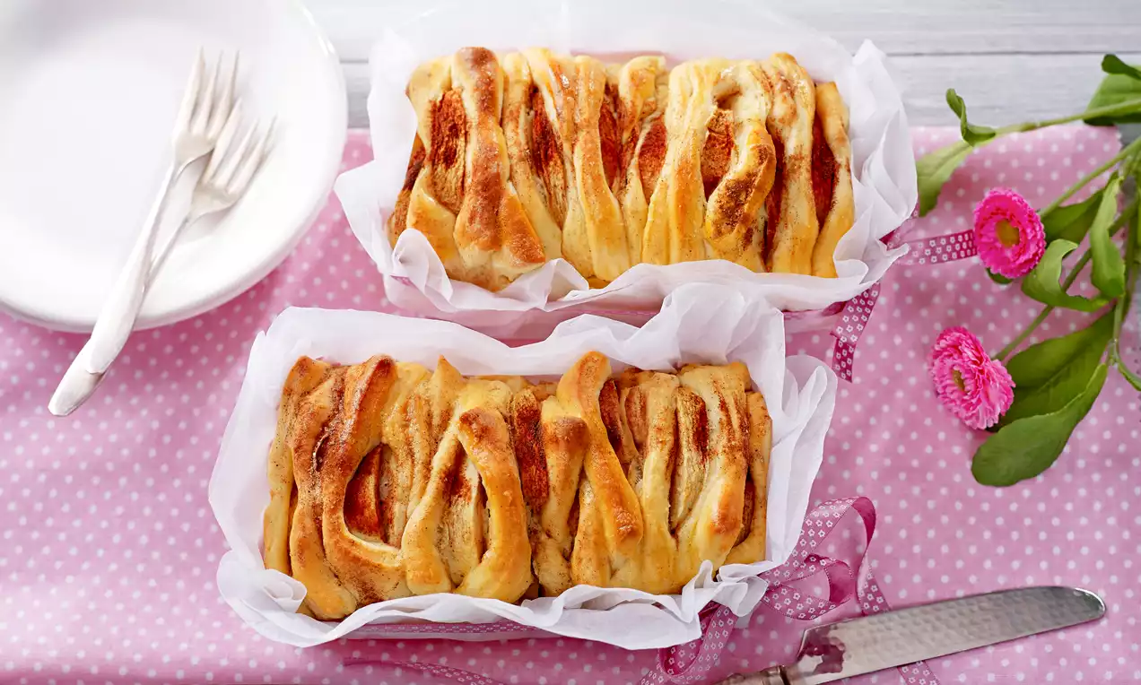 Dwa złociste chlebki drożdżowe w białym pergaminie z karbowaną fakturą, na różowej tkaninie w kropki obok talerza i kwiatów