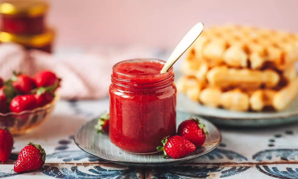 Jahodový džem sýtej červenej farby v sklenenom pohári s lyžičkou, na tanieri so štyrmi čerstvými jahodami, vedľa taniera s vaflami