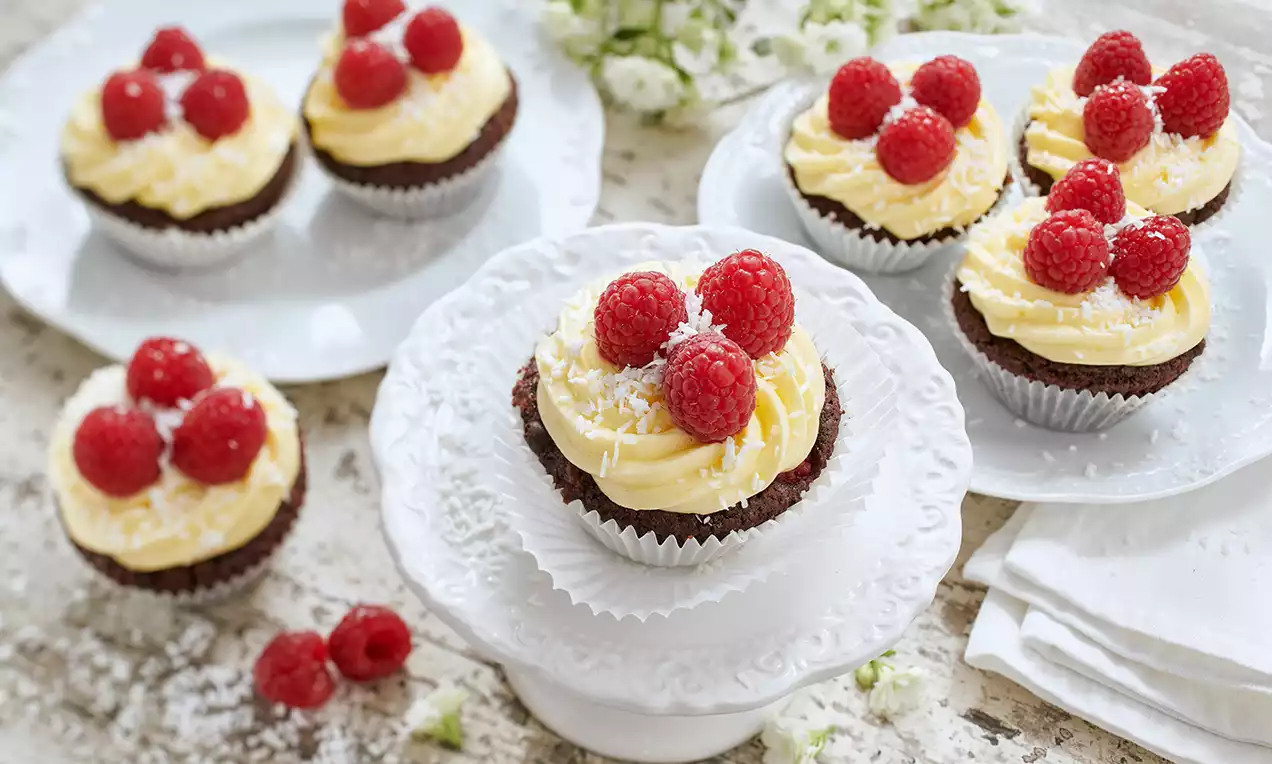 Vegane Himbeer-Kokos-Cupcakes mit gelber Creme, roten Himbeeren und weißer Kokosraspel auf weißen Tellern angerichtet