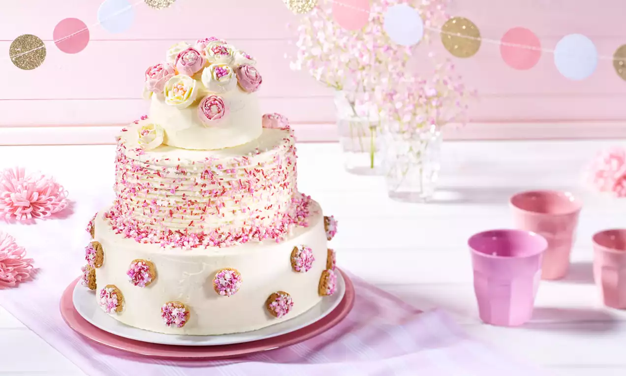 Zweistöckige Torte mit weißer Creme, rosa Streuseln und Rosendekor auf pastellfarbenem Tortenteller, umgeben von luftigem Partydekor