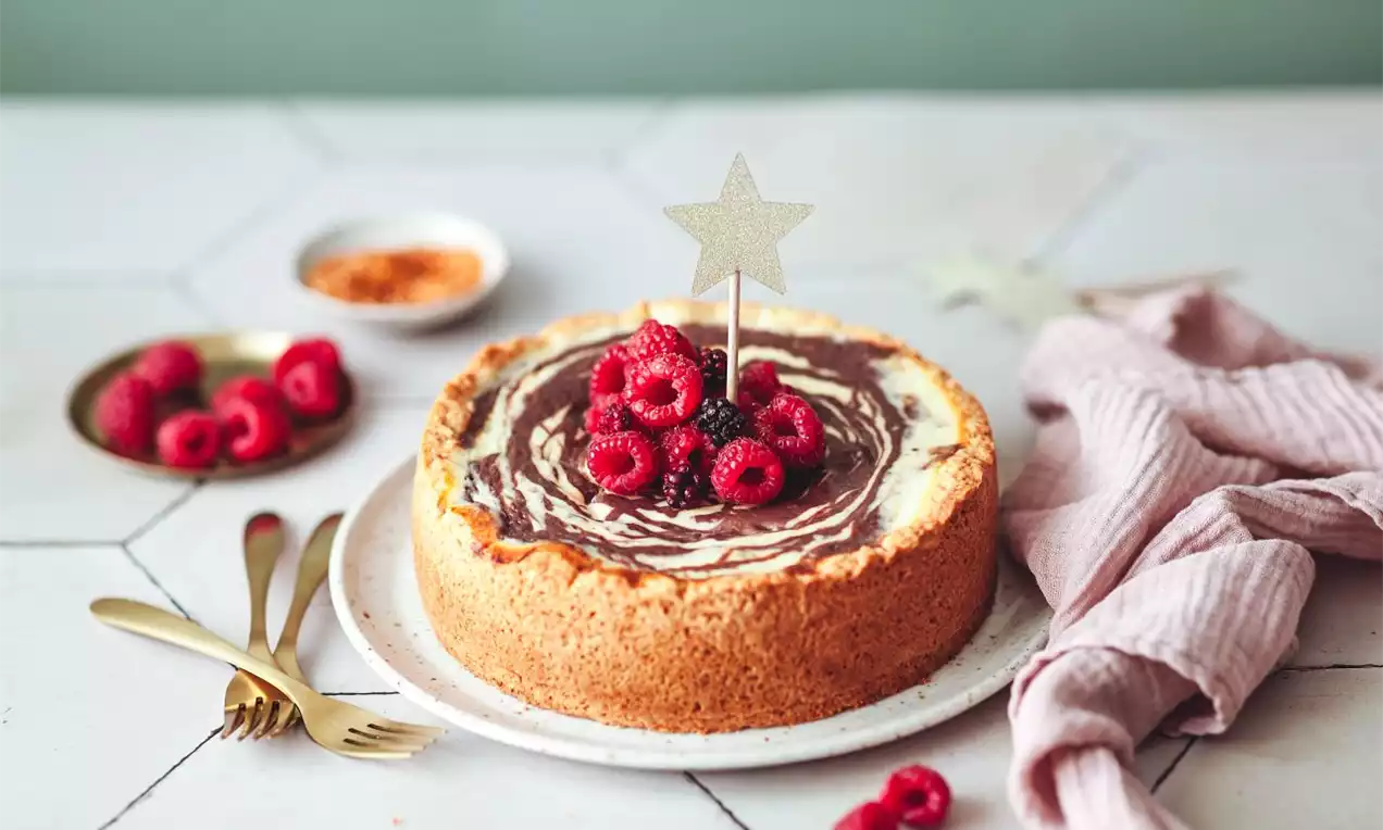 Zebra cheesecake s jemně mramorovaným krémem, zlatavou sušenkovou základnou, ozdobený čerstvými malinami a hvězdou na světlém talíři