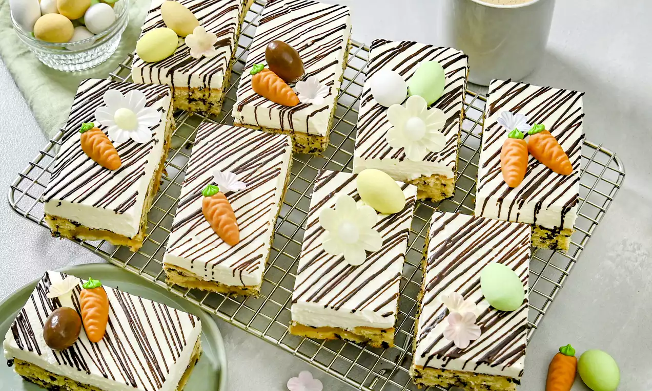 Rechteckige Kuchenstücke mit heller Creme, dunklen Schokostreifen und Dekoration aus Marzipan-Möhren, Blumen und Ostereiern auf Gitter und Teller