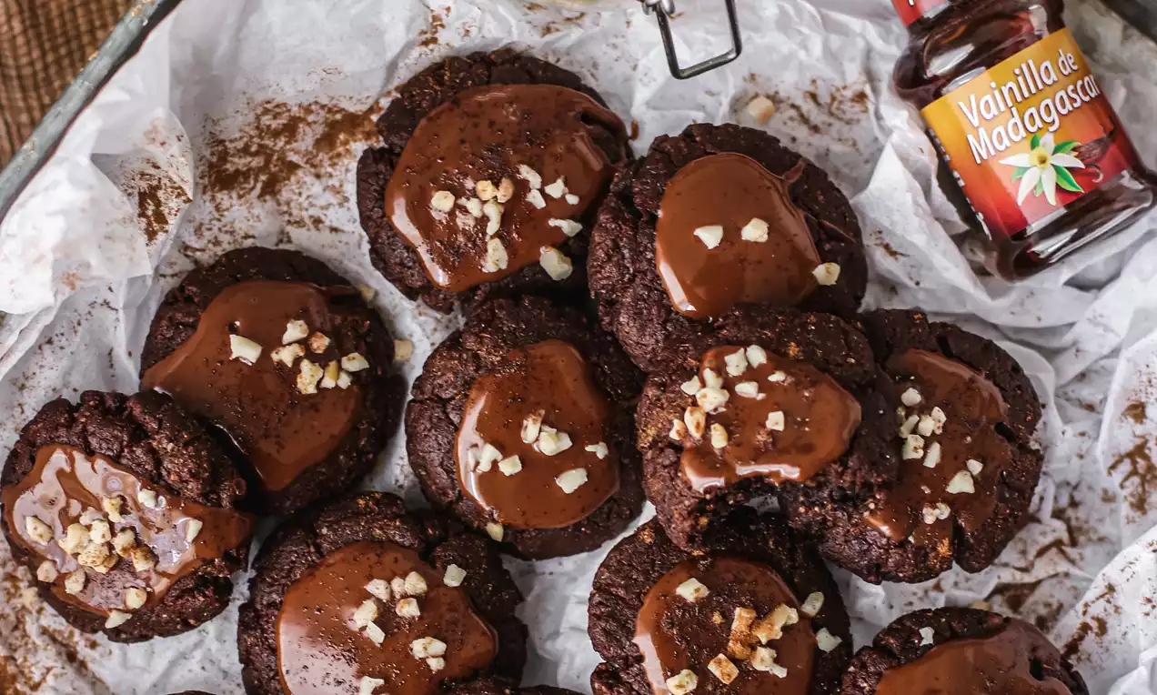 Galletas redondas de chocolate oscuro con bordes rugosos, cubiertas con glaseado brillante y trozos de almendra sobre papel encerado