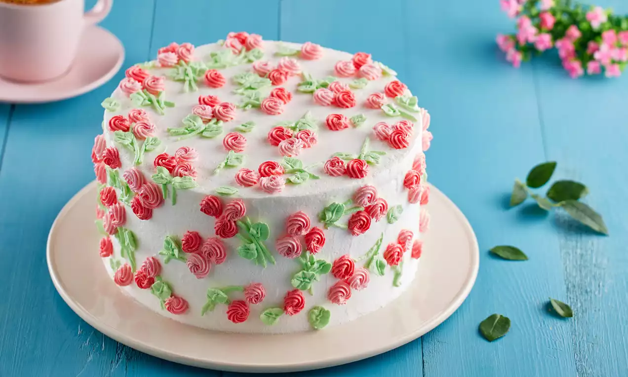 Beyaz krema kaplı pasta, pembe kremalı çiçek desenleri ve yeşil yapraklarla süslenmiş, açık mavi zeminde beyaz tabakta sunulmuş