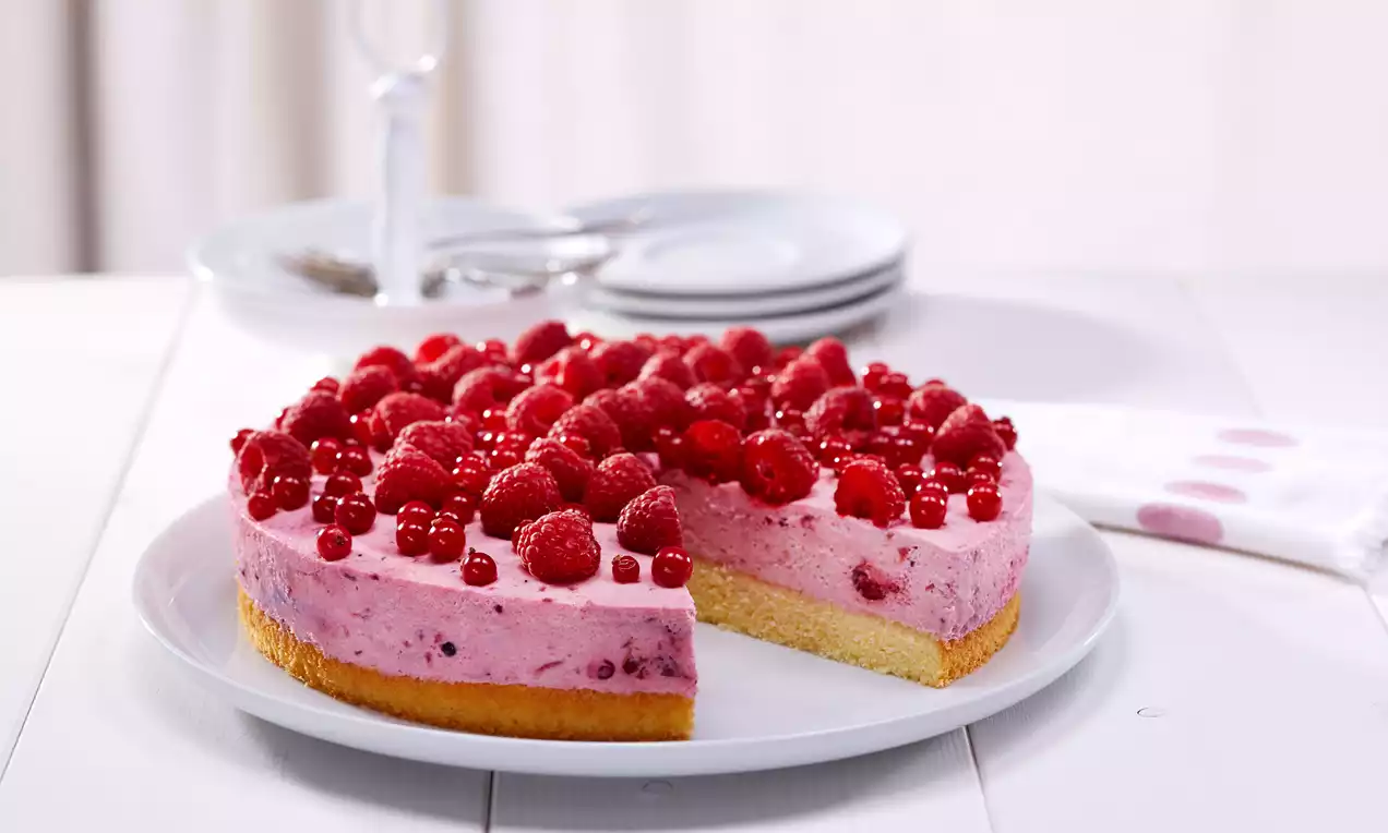 Runde Torte mit hellrosa Beerencreme auf hellem Biskuitboden, belegt mit glänzenden Himbeeren und Johannisbeeren auf weißem Teller