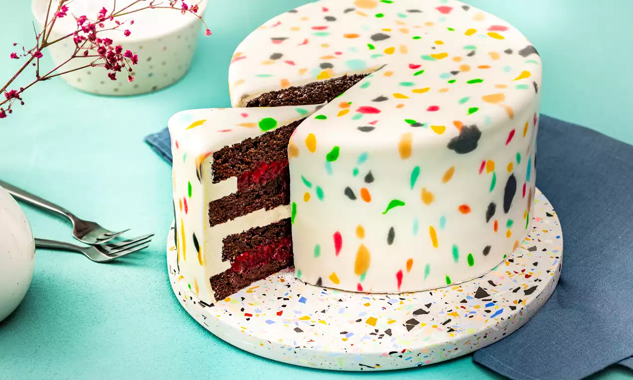 Vegaaninen Terrazzo-kakku valkoisella, kirjavilla väriroiskeilla koristellulla pinnalla, sisällä suklaakerroksia ja punaisia täytteitä, aseteltuna terrazzo-kuvioiselle tarjoilualustalle