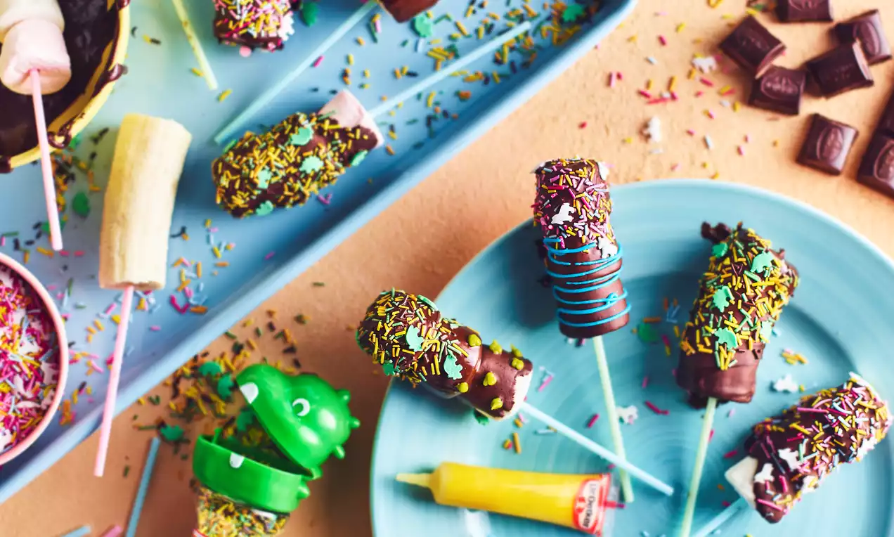 Chocolate-coated banana and marshmallow pops on sticks, sprinkled with colourful decorations, arranged on blue plates