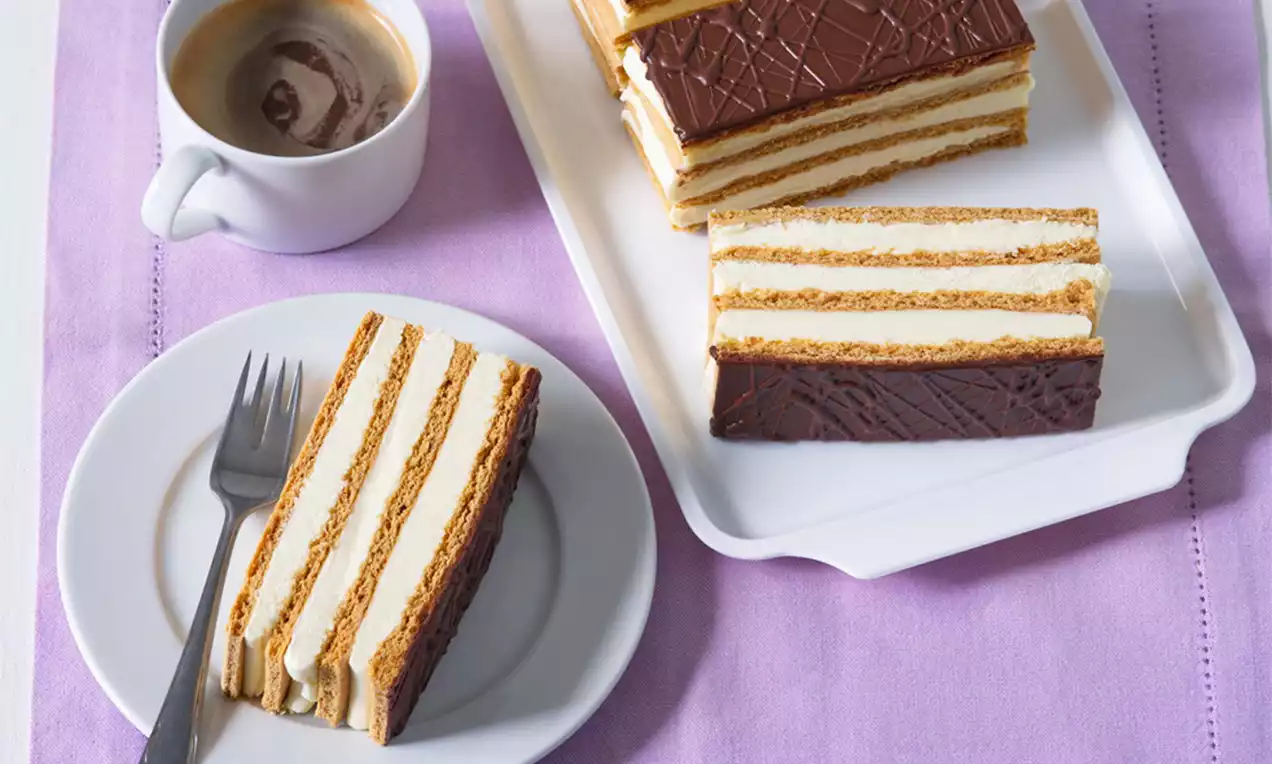 Medové rezy s jemne zlatistým cestom, vrstvami svetlej krémovej plnky a čokoládovou polevou, usporiadané na bielom tanieri s šálkou kávy