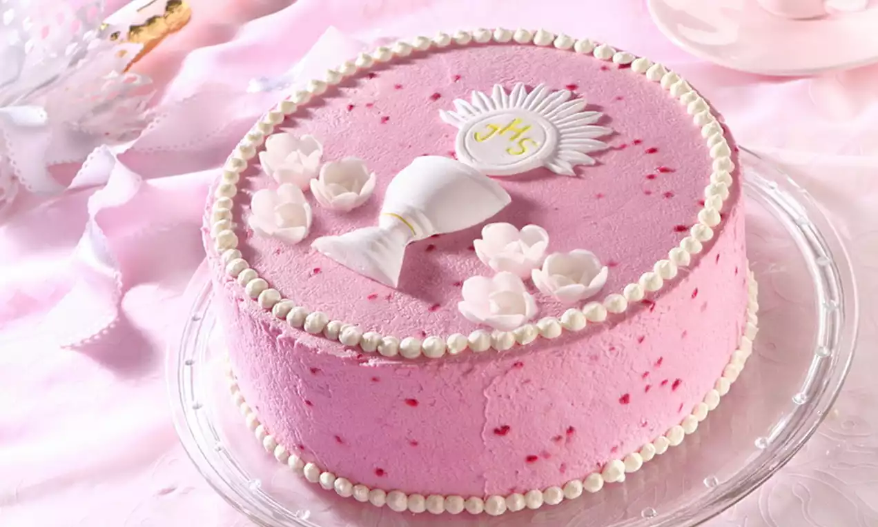 Tort o malinowym smaku w kolorze jasnoróżowym, ozdobiony perełką z kremu, białymi kwiatami i hostią na przejrzystym szklanym talerzu